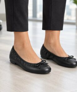 Woman wearing black vegan leather ballet flats with front bow in a bright modern office, cropped from the knees down