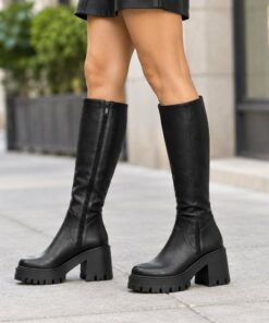 Woman wearing black vegan leather knee-high platform boots with lug sole on a city sidewalk, cropped from the thighs down
