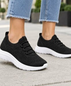Woman wearing black knit slip-on sneakers with elastic laces on a city sidewalk, cropped from the knees down