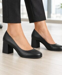 Woman wearing black vegan leather round toe pumps with low 2 inch block heel in a bright modern office, cropped from the knees down
