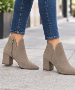 Woman wearing taupe suede-look V-cut ankle booties with low block heel on a city sidewalk, cropped from the knees down