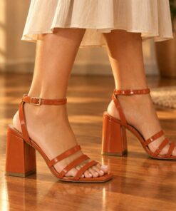 Woman wearing terracotta patent strappy sandals with 2.5 inch block heel at an evening party, cropped from the knees down